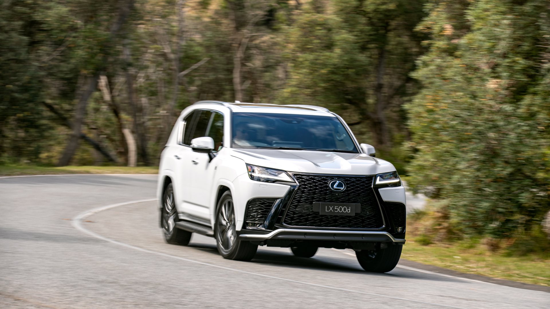 A white Lexus LX turns left around a corner on a highway.