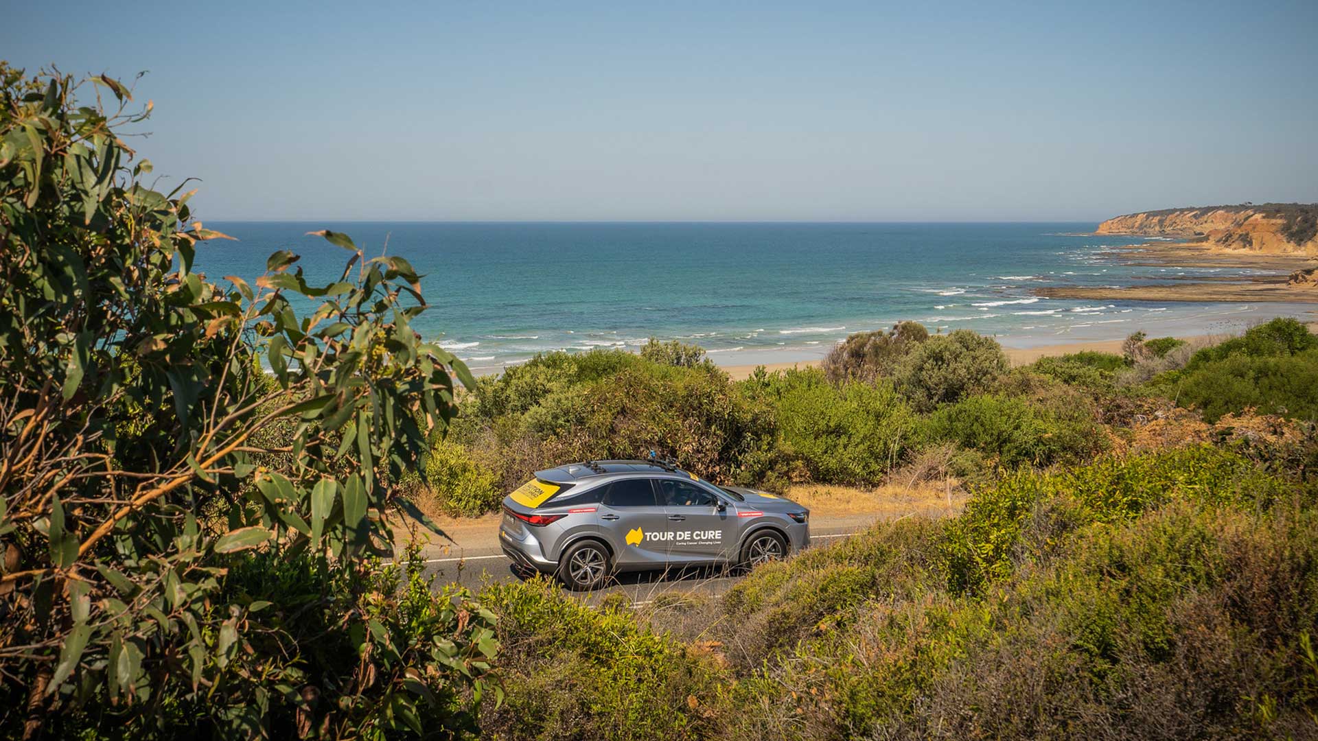 A Lexus Tour de Cure support vehicle drives along a coastal road overlooking the ocean.