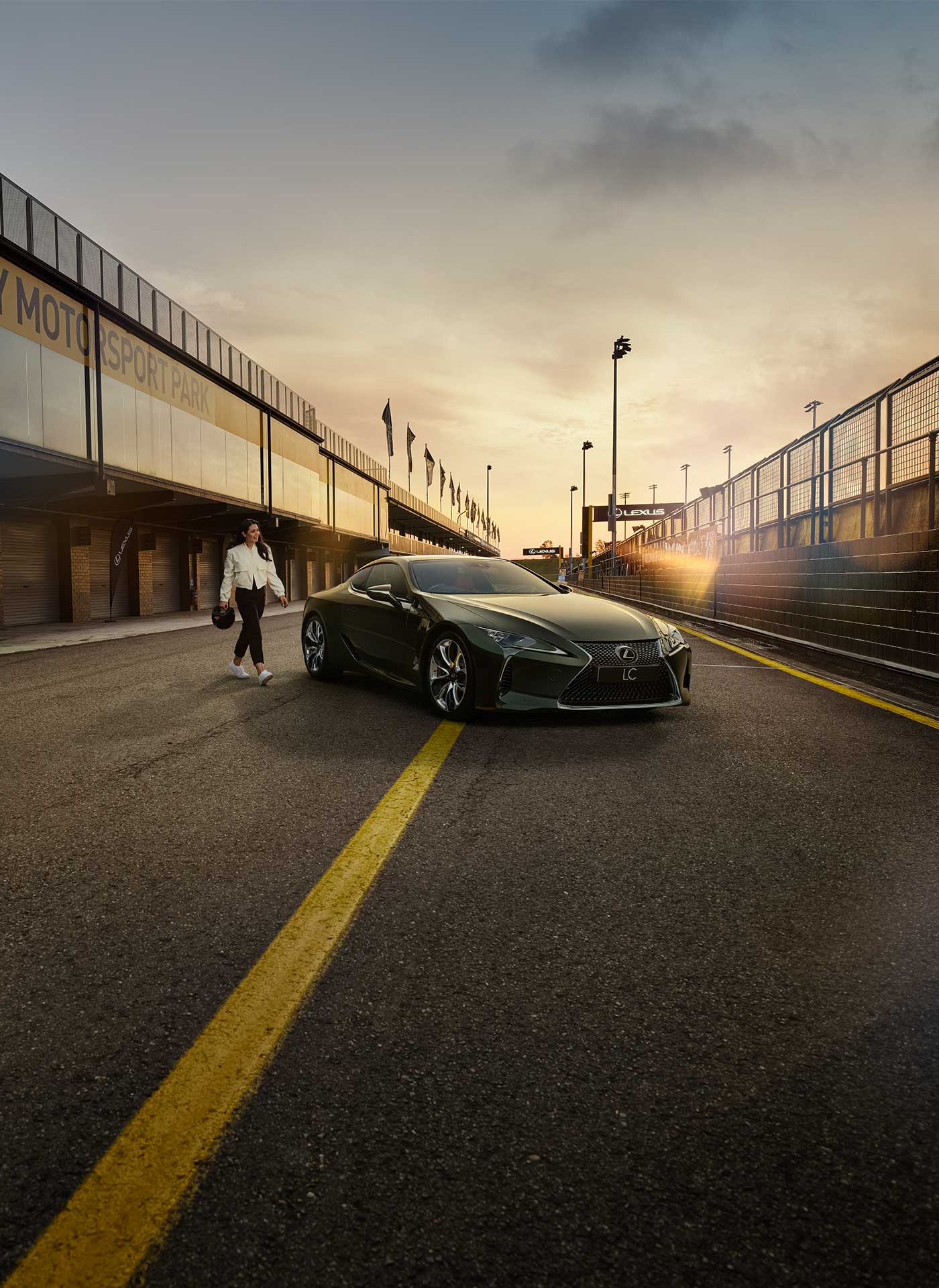 A woman dressed in a white jacket and black pants and holding a helmet walks towards an LC coupe parked in a pit lane